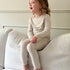 Mini Muse PJs — Dusk: Adorable toddler in beige long-sleeve pajamas sitting on a couch.